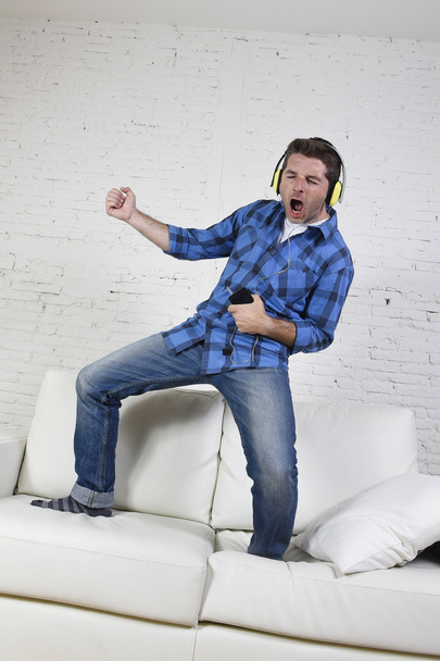 20s or 30s man jumped on couch listening to music on mobile phone with headphones playing air guitar - Foto, Imagem