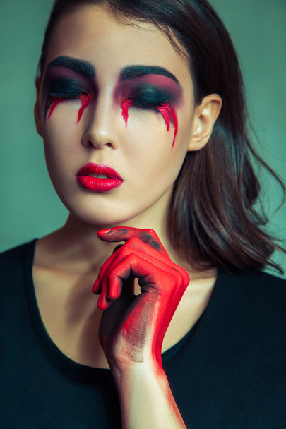 portrait of freak monster with mess dirty colored makeup on her face. crying woman with red bloody tears and hand. halloween concept on green background. studio shot, dark brown eyes. - 写真・画像