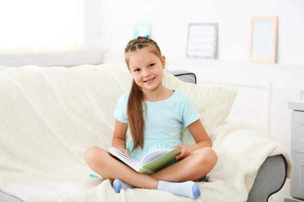 Маленька дівчинка сидить на дивані з книгою
  - Фото, зображення
