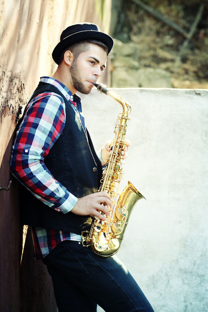 Young man playing on saxophone outside near the old wall - 写真・画像
