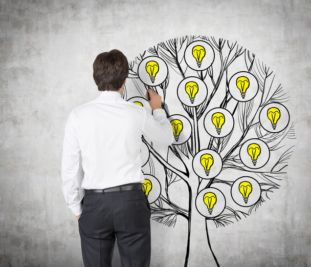 Rückansicht eines jungen Profis, der einen Baum mit Glühbirnen an die Betonwand zeichnet. Glühbirnen als Konzept für neue Geschäftsideen. - Foto, Bild
