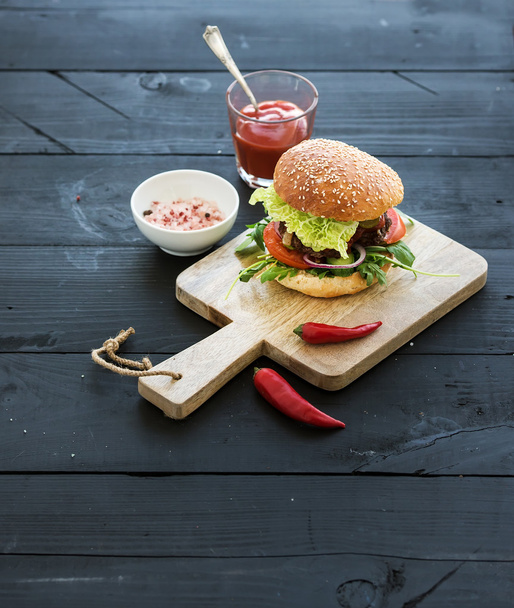 Fresh homemade burger on dark serving board with spicy tomato sauce, sea salt and herbs over black wooden background. Top view. - Fotó, kép