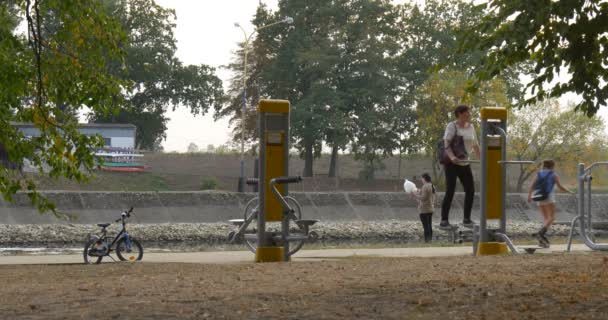 Sport grond Speeltuin vrouw doet oefeningen twee meisjes op rolschaatsen mensen lopen met hond kind in fietshelm geparkeerd blauwe fiets groene bomen - Video
