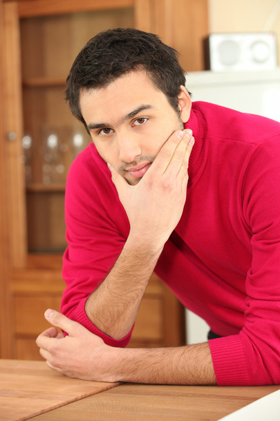 Young dark-haired guy in kitchen wearing raspberry red jumper - 写真・画像