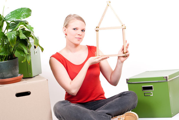 Young woman with house symbol sits between move cardboards and plant - Photo, Image