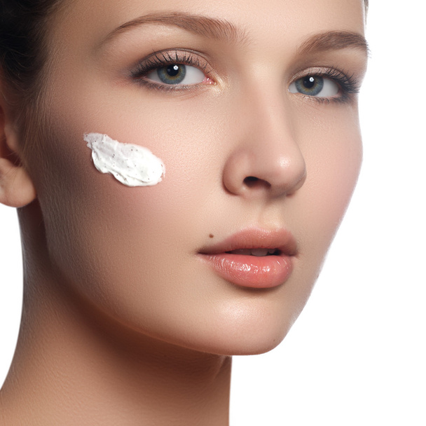 Beautiful face of young woman with cosmetic cream on a cheek. Skin care concept. Closeup portrait isolated on white. Close-up young beautiful face of girl applying moisturize cream - Zdjęcie, obraz
