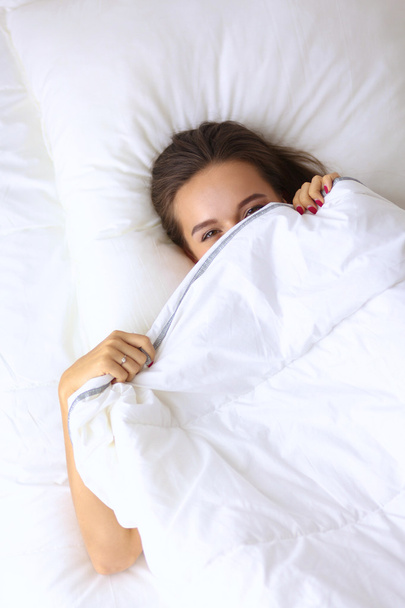 A pretty young woman peeking from under the covers in her bedroom happilly - 写真・画像