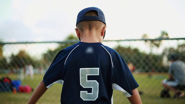 Маленька дитина в бейсбольній формі спостерігає за практикою за парканом на полі
 - Кадри, відео