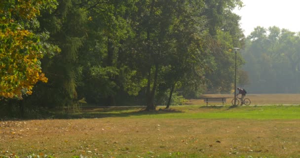 公園のベンチの近くで自転車を持つ男は、彼は自転車に乗り、ベージュのパンツとマルーンシャツ晴れた秋の日に男を離れて乗ります - 映像、動画