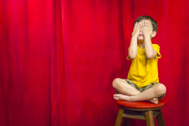 Niño cubriendo los ojos sentado en el taburete delante de la cortina
 - Foto, Imagen