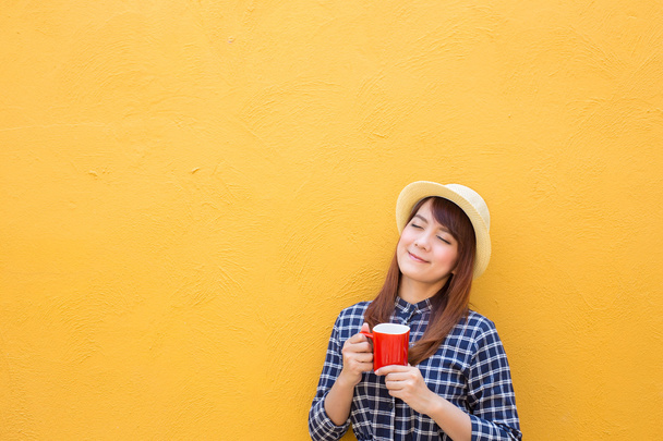 smiling woman wear in dress and hat holding red coffee cup on yellow cement wall background, thinking, imagination - Φωτογραφία, εικόνα