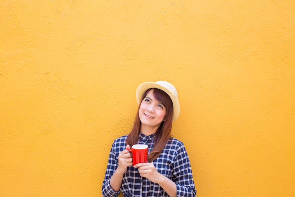 smiling woman wear in dress and hat holding red coffee cup on yellow cement wall background, thinking, imagination - Fotoğraf, Görsel