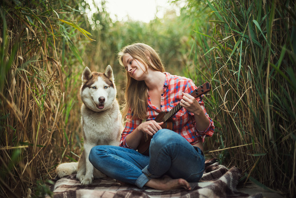 young caucasian woman playing ukulele. Female with siberian husky dog playing guitar outdoors - 写真・画像