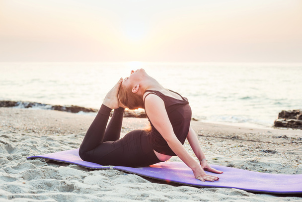 young caucasian female practicing yoga on beach during  sunrise - Photo, Image