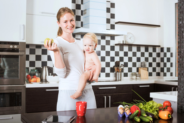 Mother Holding Daughter In Kitchen while cooking a meal of fresh vegetables and fruits. Mother and little daughter on kitchen holding an apple. Healthy food and healthy living concept - Fotoğraf, Görsel