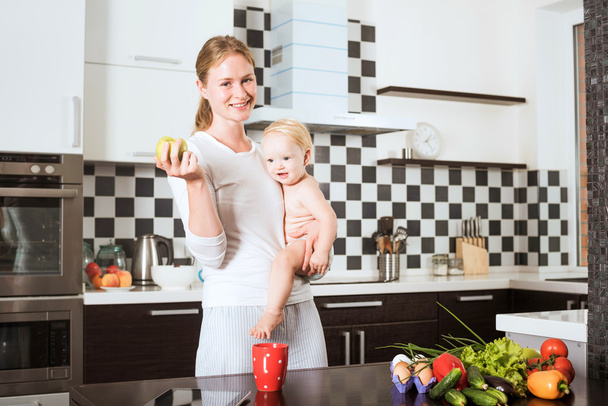 Anne Holding kızı taze sebze ve meyve yemek sırasında mutfak. Anne ve küçük kızı bir elma tutan mutfak üzerinde. Sağlıklı gıda ve sağlıklı yaşam kavramı - Fotoğraf, Görsel