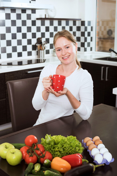 young caucasian female drinking coffee or tea in kitchen, fresh fruits and vegetables on table. She starts her morning with healthy breakfast - Foto, Imagem