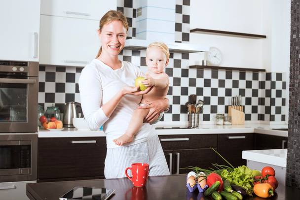 Mother Holding Daughter In Kitchen while cooking a meal of fresh vegetables and fruits. Mother and little daughter on kitchen holding an apple. Healthy food and healthy living concept - Foto, afbeelding