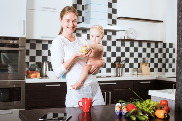 Mother Holding Daughter In Kitchen while cooking a meal of fresh vegetables and fruits. Mother and little daughter on kitchen holding an apple. Healthy food and healthy living concept - Fotoğraf, Görsel