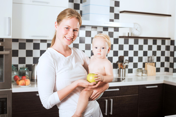 Mother Holding Daughter In Kitchen while cooking a meal of fresh vegetables and fruits. Mother and little daughter on kitchen holding an apple. Healthy food and healthy living concept - Zdjęcie, obraz