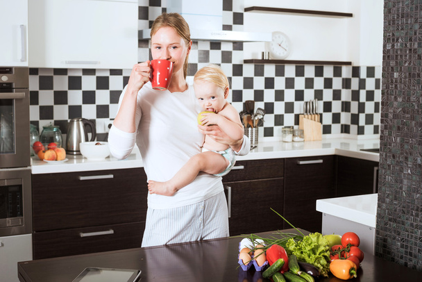 Mother Holding Daughter In Kitchen while cooking a meal of fresh vegetables and fruits. Mother and little daughter on kitchen holding an apple. Healthy food and healthy living concept - Foto, Imagem
