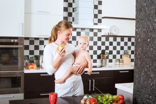 Mother Holding Daughter In Kitchen while cooking a meal of fresh vegetables and fruits. Mother and little daughter on kitchen holding an apple. Healthy food and healthy living concept - Foto, immagini