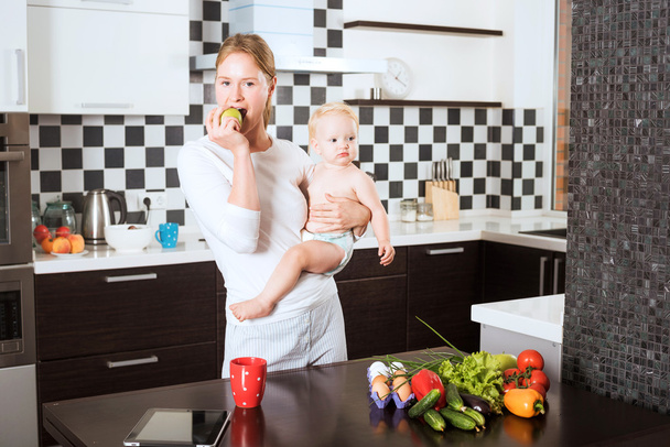 Mother Holding Daughter In Kitchen while cooking a meal of fresh vegetables and fruits. Mother and little daughter on kitchen holding an apple. Healthy food and healthy living concept - Фото, изображение