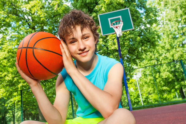 Happy boy sits on playground, holds ball near face - Фото, изображение