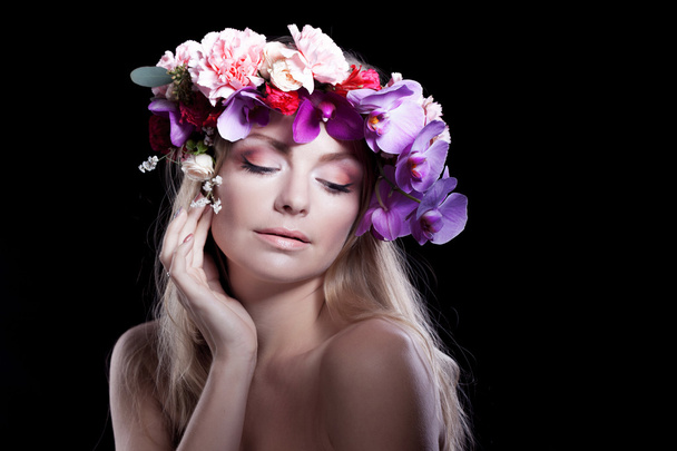 young beautiful woman in wreath of flowers holds hand to face, black background - Фото, изображение