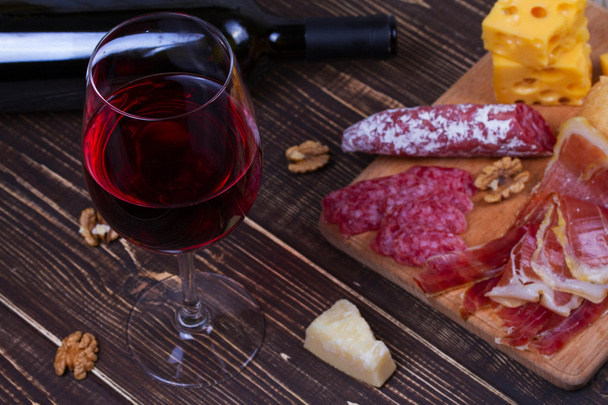 Copo e garrafa de vinho tinto, queijo, salame, nozes, prosciutto e alecrim sobre fundo de madeira
 - Foto, Imagem