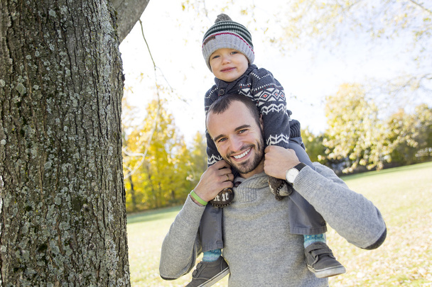 Portrait of happy father giving son piggyback ride on his shoulders in autumn park. - Фото, изображение
