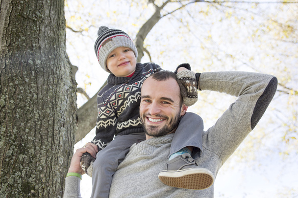 Portrait of happy father giving son piggyback ride on his shoulders in autumn park. - Photo, image