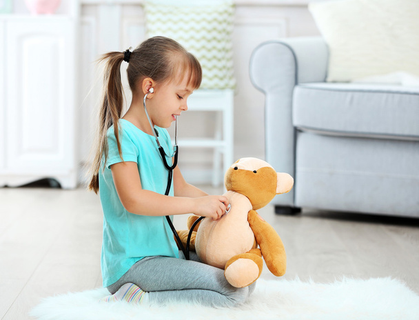 Schattig meisje met stethoscoop en teddy bear zittend op tapijt, op huis interieur achtergrond - Foto, afbeelding