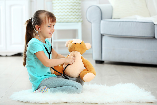 Little cute girl with stethoscope and teddy bear sitting on carpet, on home interior background - 写真・画像