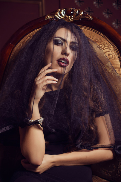 Mysterious young beautiful woman in black transparent lace veil, evening dress sitting in vintage chair. Accessories pearl bracelet. Fashion make-up smoky eyes. Black and white shades. luxury. Sexuality. Seduction. Sensuality. - Photo, Image