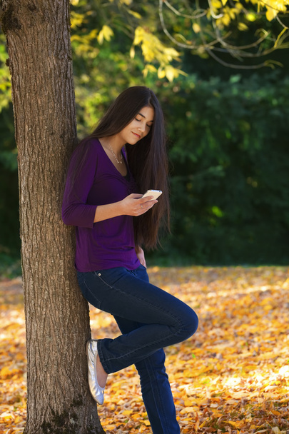 Teen girl standing against autumn tree looking at cell phone - Foto, Bild