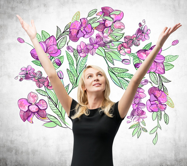 Een verbaasd blonde vrouw met handen omhoog en ze droomt over zomerbloemen. De schets van paarse bloemen wordt op de betonnen muur getekend. - Foto, afbeelding