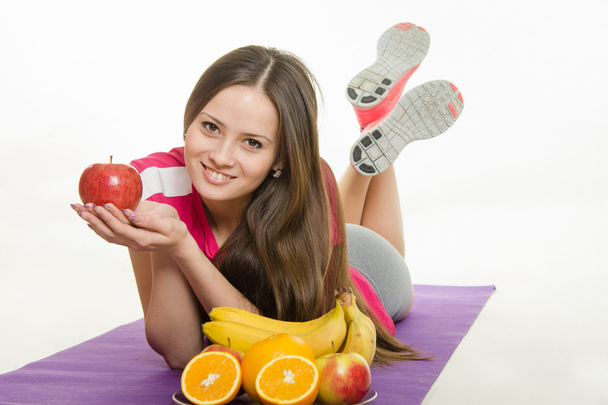 Sportswoman couché sur un tapis, tenant une pomme à la main, il est confronté à une assiette de fruits
 - Photo, image