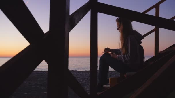 Ταξίδια: σιλουέτα όμορφο κορίτσι τουρίστα που ζωγραφίζει το ηλιοβασίλεμα στην παραλία. Μεσαία βολή, αργή κίνηση 60fps, στατική. - Πλάνα, βίντεο