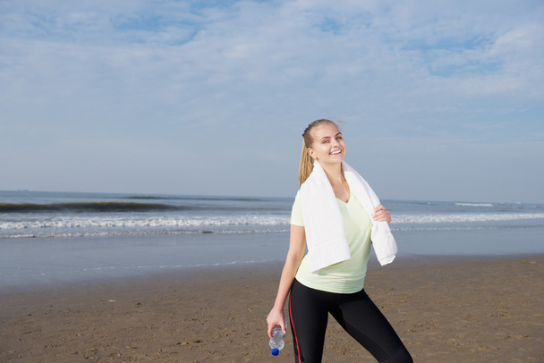 Улыбающаяся спортивная женщина, стоящая с бутылкой воды на пляже
 - Фото, изображение