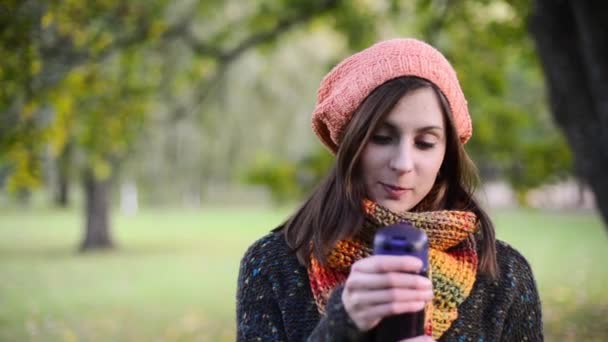 Beautiful young woman opens a thermocup and drink hot tea from it  in autumn park. - Záběry, video