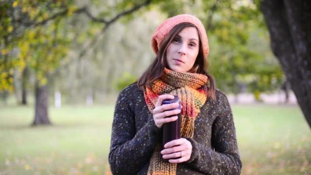 Beautiful young woman opens a thermocup and drink hot tea from it in autumn park. - Video