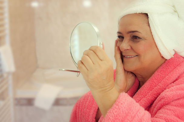Portrait of an attractive middle aged woman looking into a mirror in the bathroom - Foto, immagini