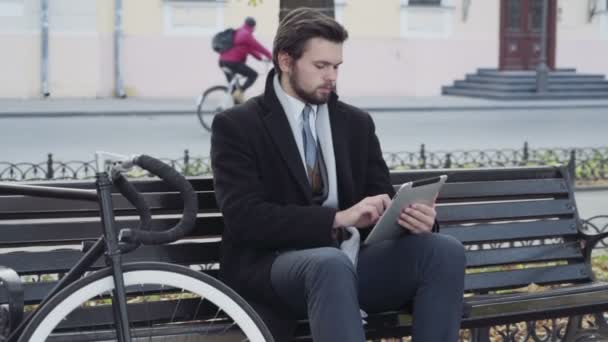 businessman using a tablet sitting on a bench near the bicycle - Imágenes, Vídeo