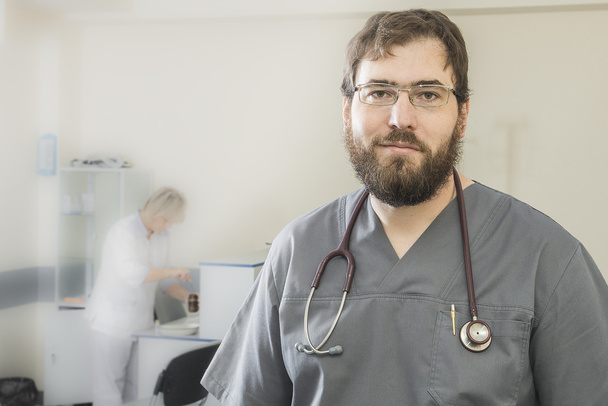 bearded doctor wearing glasses and a gray robe stands in the foreground to the background to blur assistant - Photo, Image