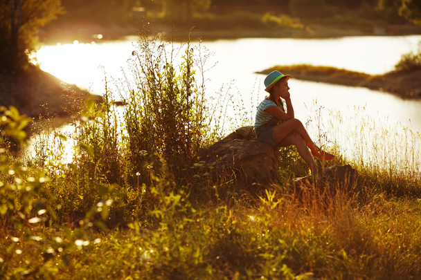 Girl in hat sitting on the bank of the river - Φωτογραφία, εικόνα