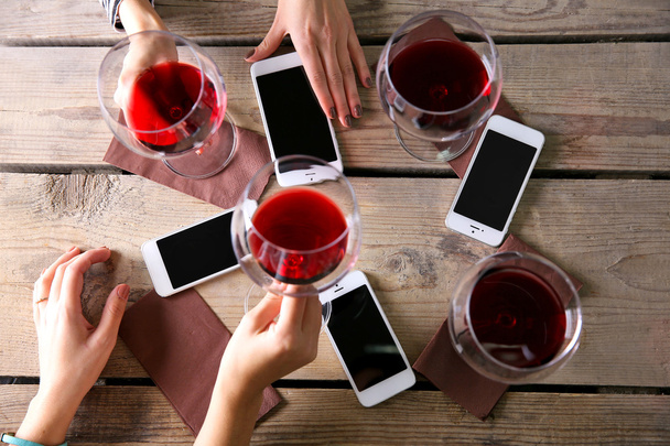 Four hands with smart phones holding glasses with red wine, on wooden table background - Фото, зображення