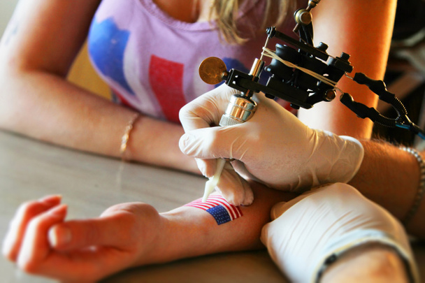 Tätowierer zeigt Prozess der Herstellung eines Tattoos auf junge schöne Hipster-Frau mit blonden Haaren Hand. Tätowierung in Form eines Pin-up-Girls mit Blütenkopf - Foto, Bild