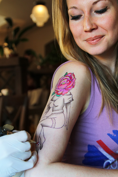 tattooer showing process of making a tattoo on young beautiful hipster woman with blonde hair hand. Tattoo design in the form pin-up girl with flower head - Photo, Image