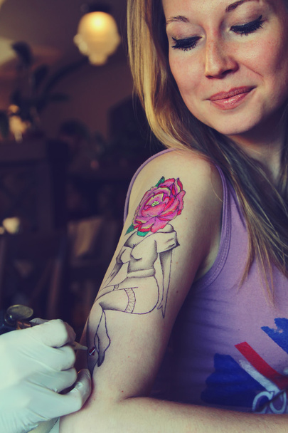 татуировщик, показывающий процесс создания татуировки на молодой красивой хипстерской женщине с белокурой волосатой рукой. Татуировка дизайн в форме пин-ап девушка с цветочной головой
 - Фото, изображение